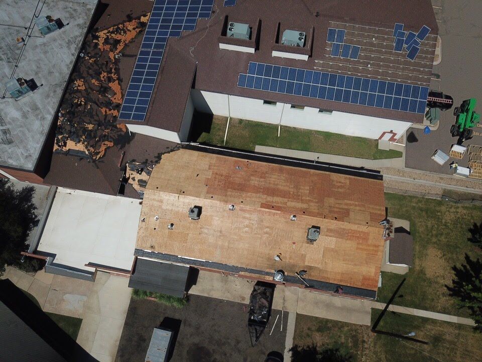 Aerial view of a building with a damaged roof and solar panels; construction materials are visible.