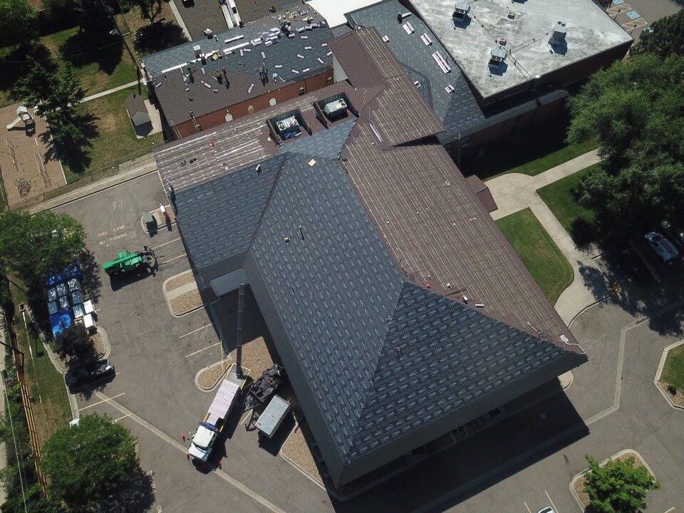Aerial view of a building with gray and brown shingled roofs, parked vehicles, and surrounding trees.