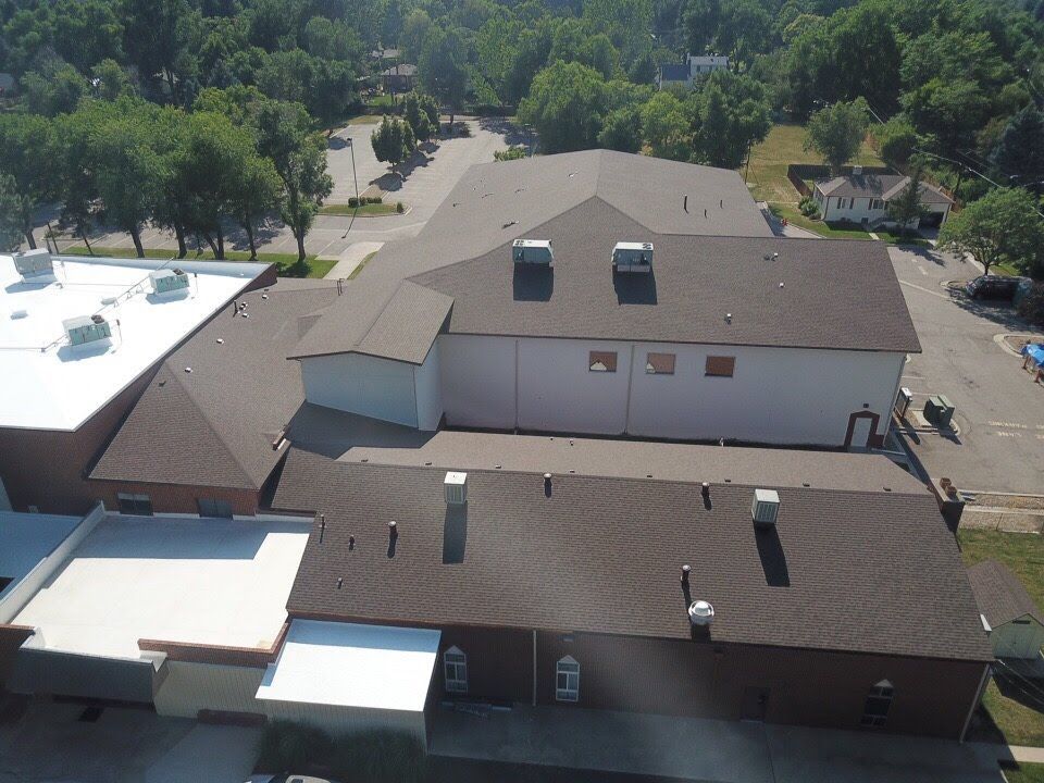 Aerial view of a brown-roofed building with a white side, surrounded by trees and a parking lot.