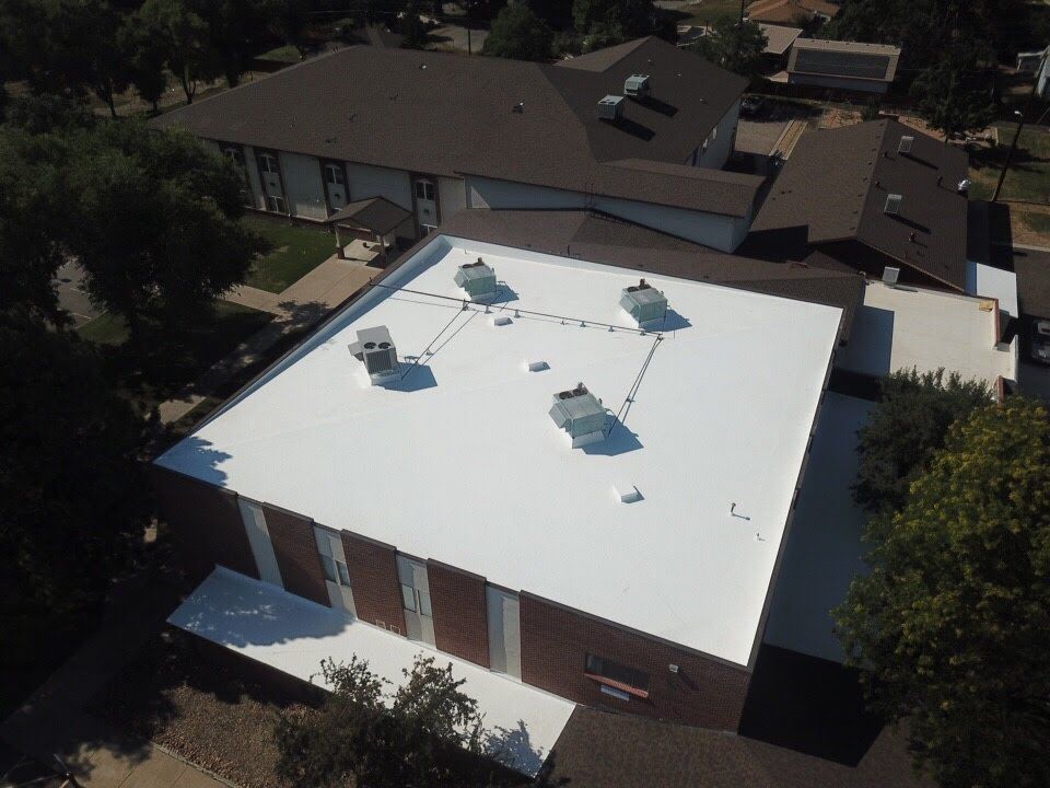 Aerial view of a white flat commercial roof with HVAC units. Red brick building, other roofs, and trees in the background.
