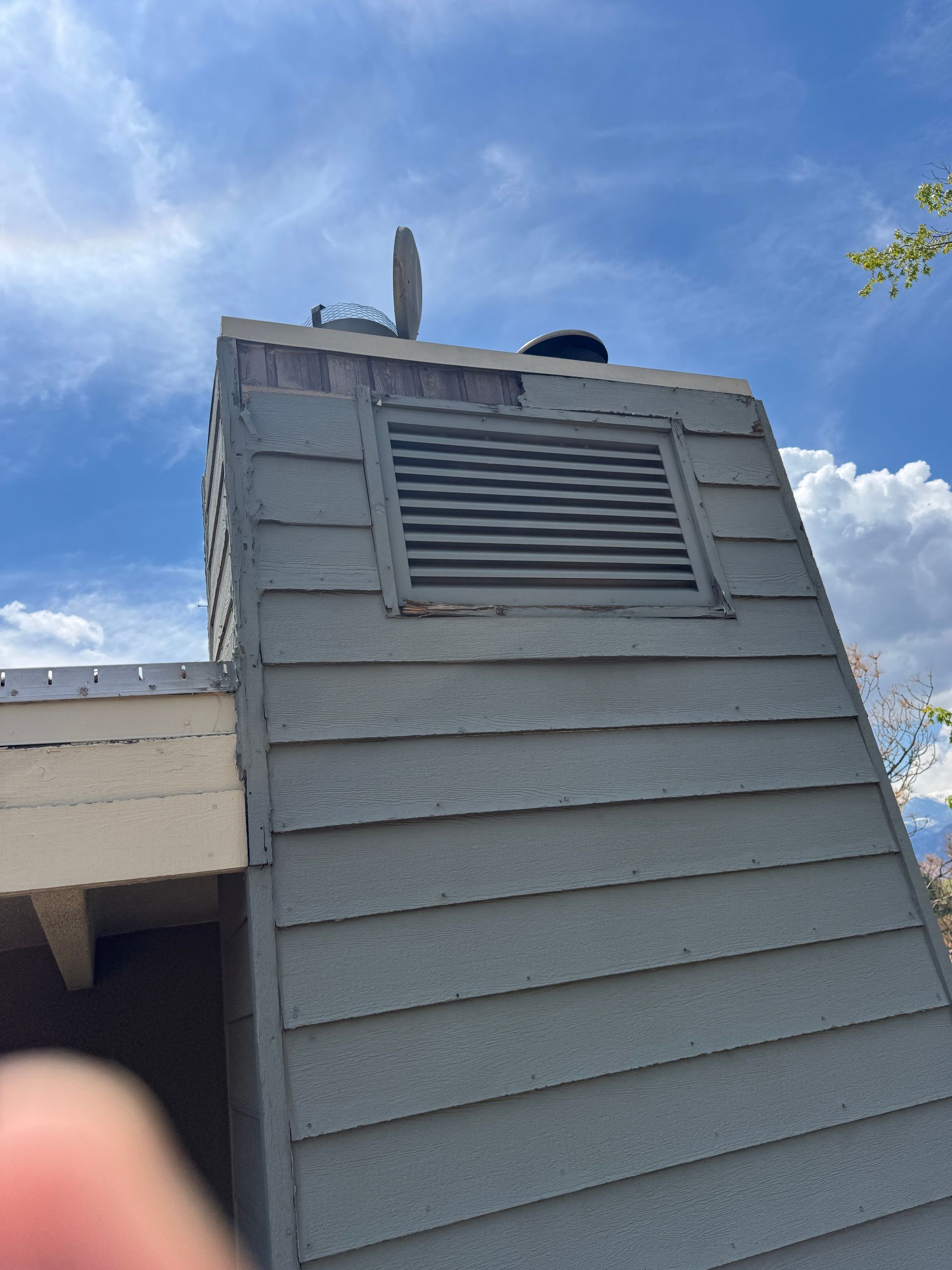 Gray chimney with louvered vent, against a blue sky with scattered clouds.