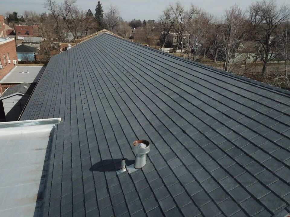 Dark gray tile roof of a building with a chimney, trees, and other buildings visible in the background.