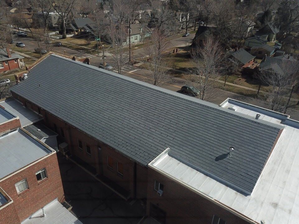 Aerial view of a long, dark gray roof on a brick building surrounded by trees and houses.
