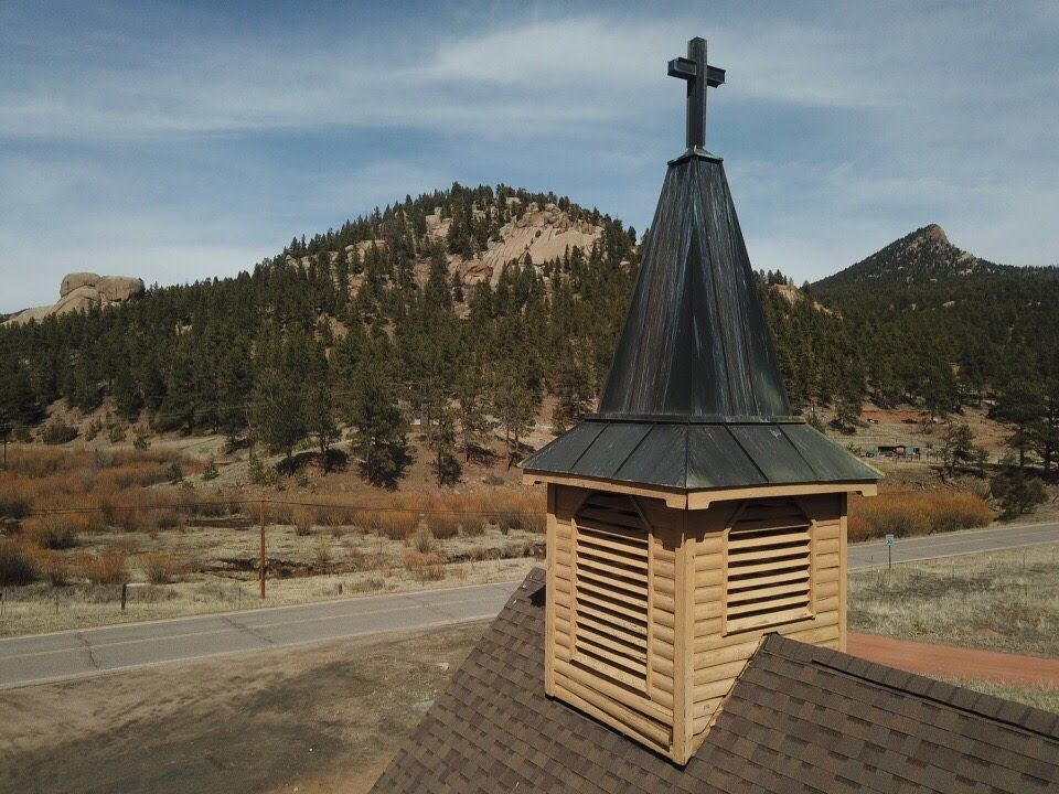 Church steeple with cross against mountain backdrop and blue sky.