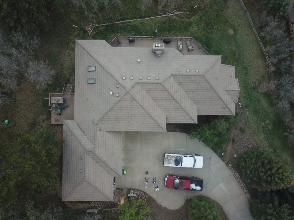 Aerial view of a house with a brown tile roof, driveway with parked cars, and surrounding trees.