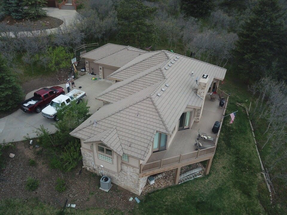 Aerial view of a house with a brown tiled roof, deck, and driveway with parked trucks, surrounded by trees.
