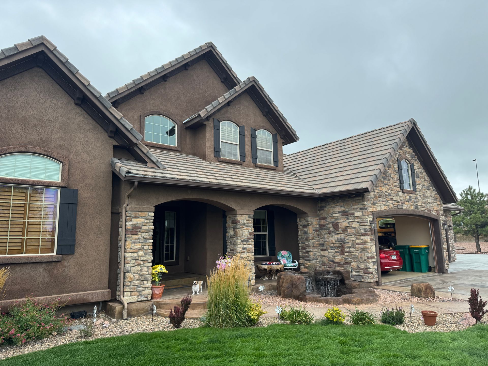 Brown stucco and stone house with arched entry, garage, and landscaping under a cloudy sky.