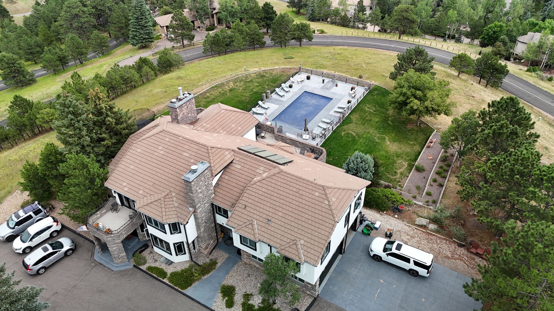 Aerial view of a large house with a pool, vehicles parked in front, and trees surrounding it.