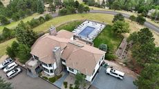 Aerial view of a large house with a pool, vehicles parked in front, and trees surrounding it.