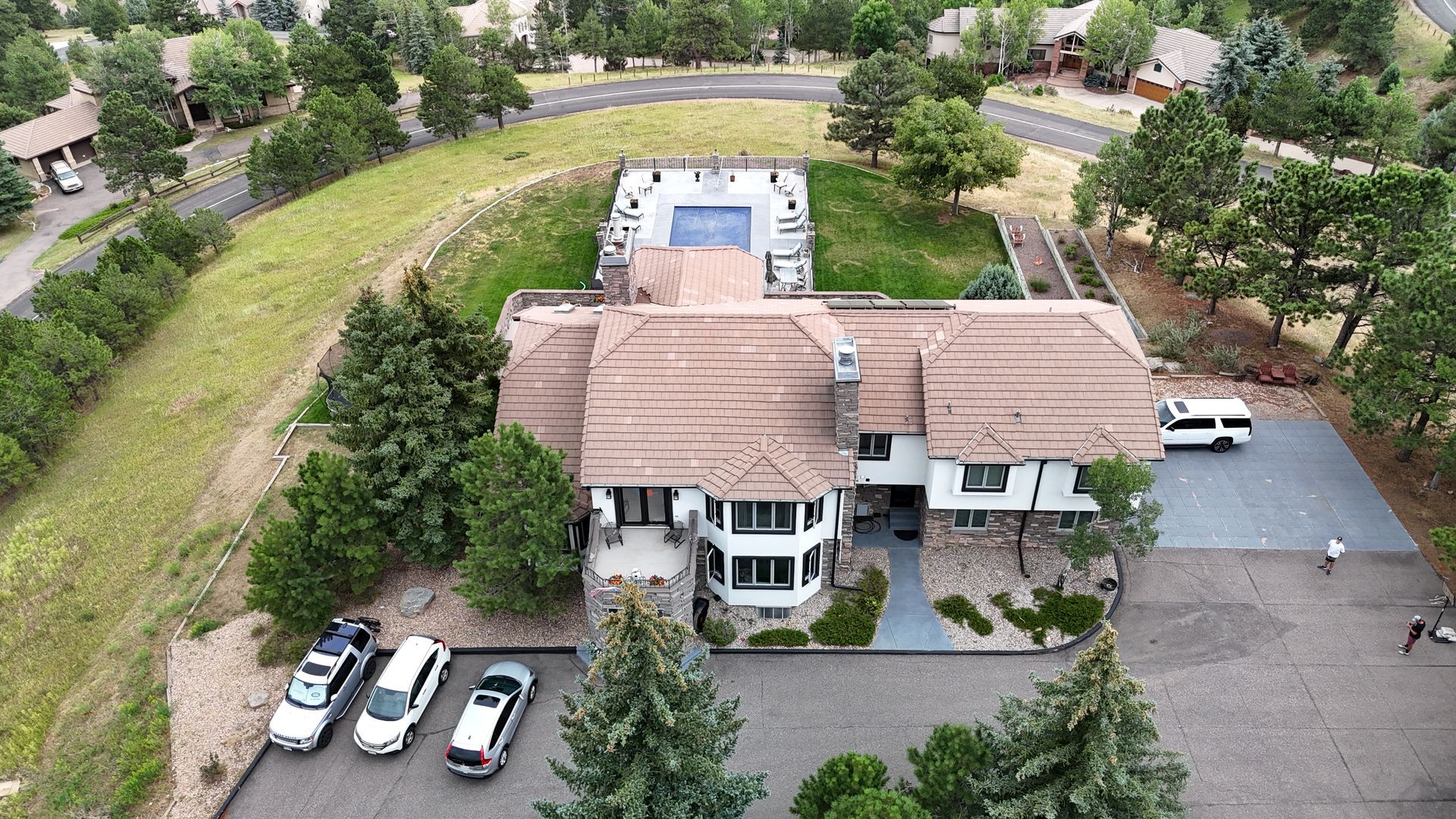 Aerial view of a large house with a pool. Cars parked in front, trees surround it.