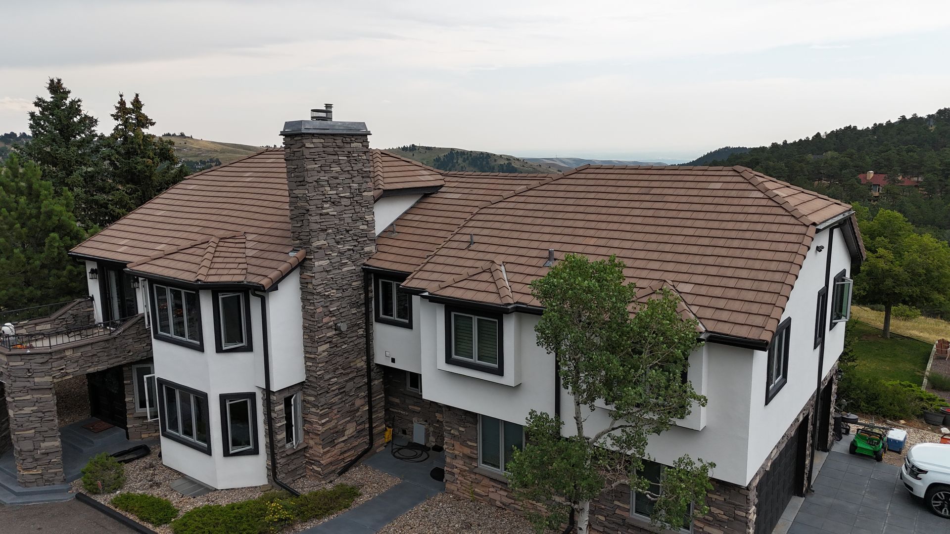 Large white house with a brown roof, stone chimney, and dark window trim, set against a green hillside.