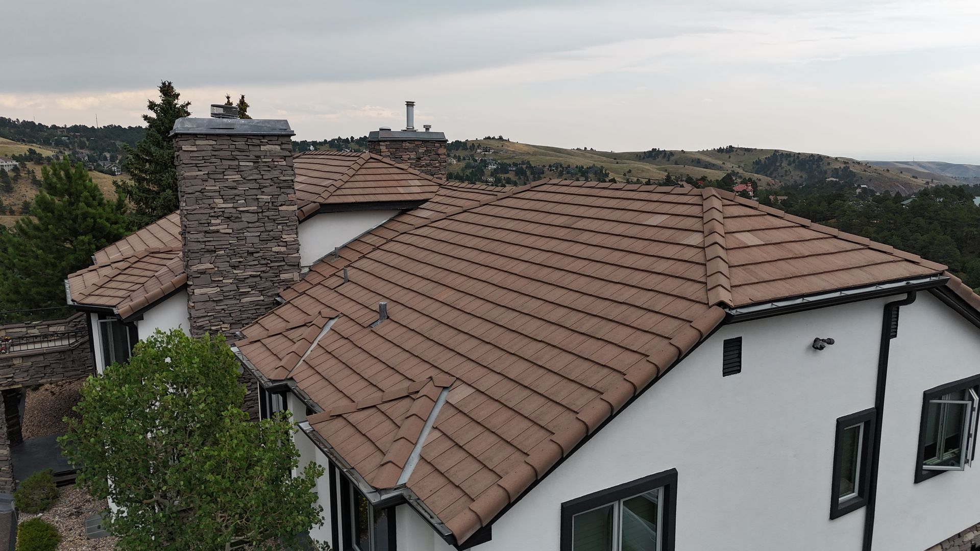 House with brown tile roof, stone chimney, white stucco walls, and dark window frames, set on a hillside.
