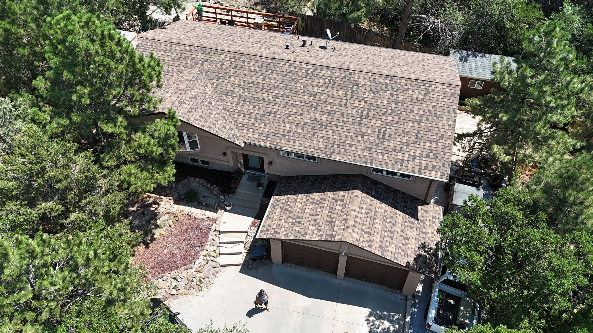 Aerial view of a house with a brown roof surrounded by green trees, driveway, and garage.