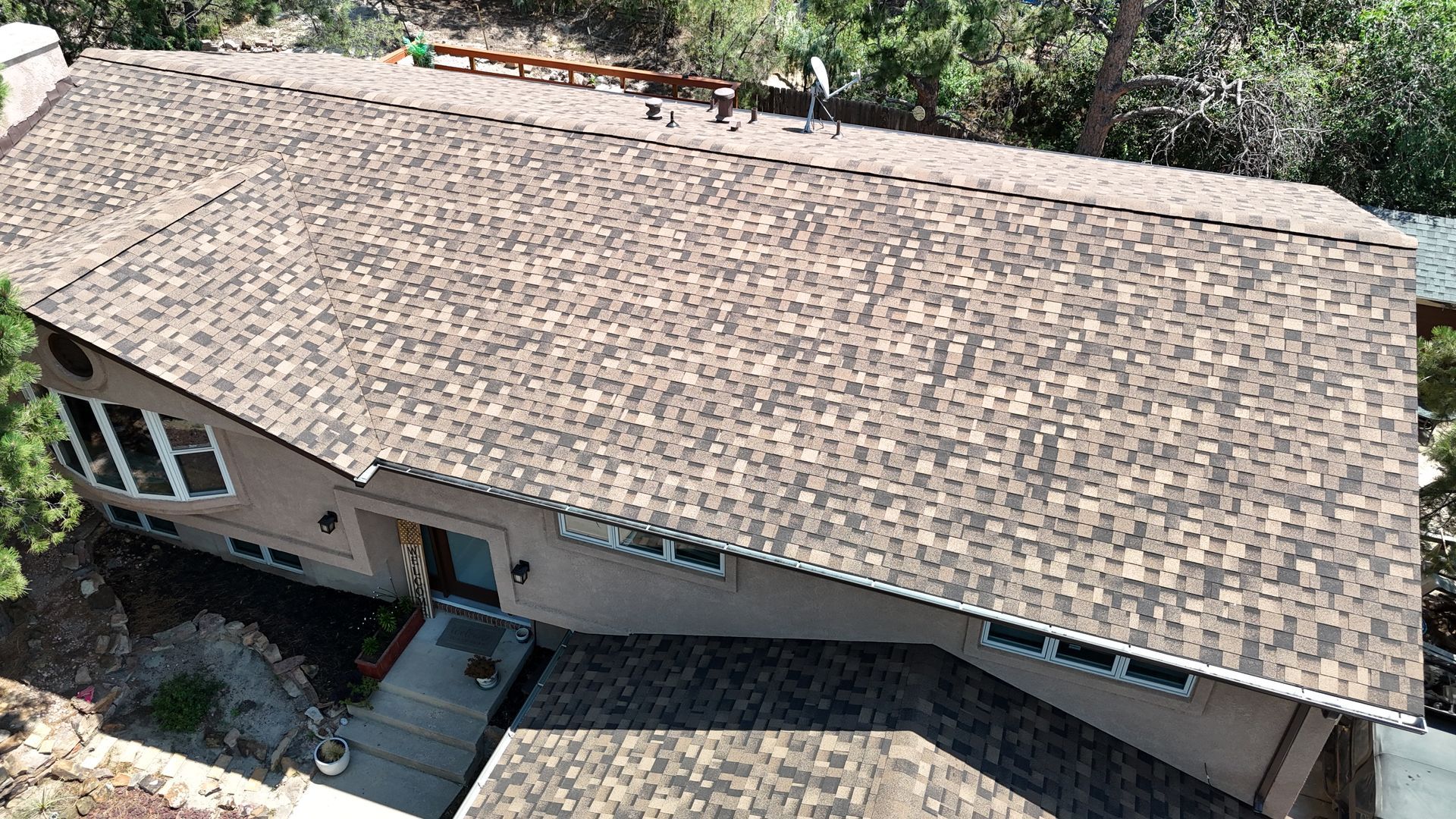 Aerial view of a house with a brown shingled roof, surrounded by trees and greenery.