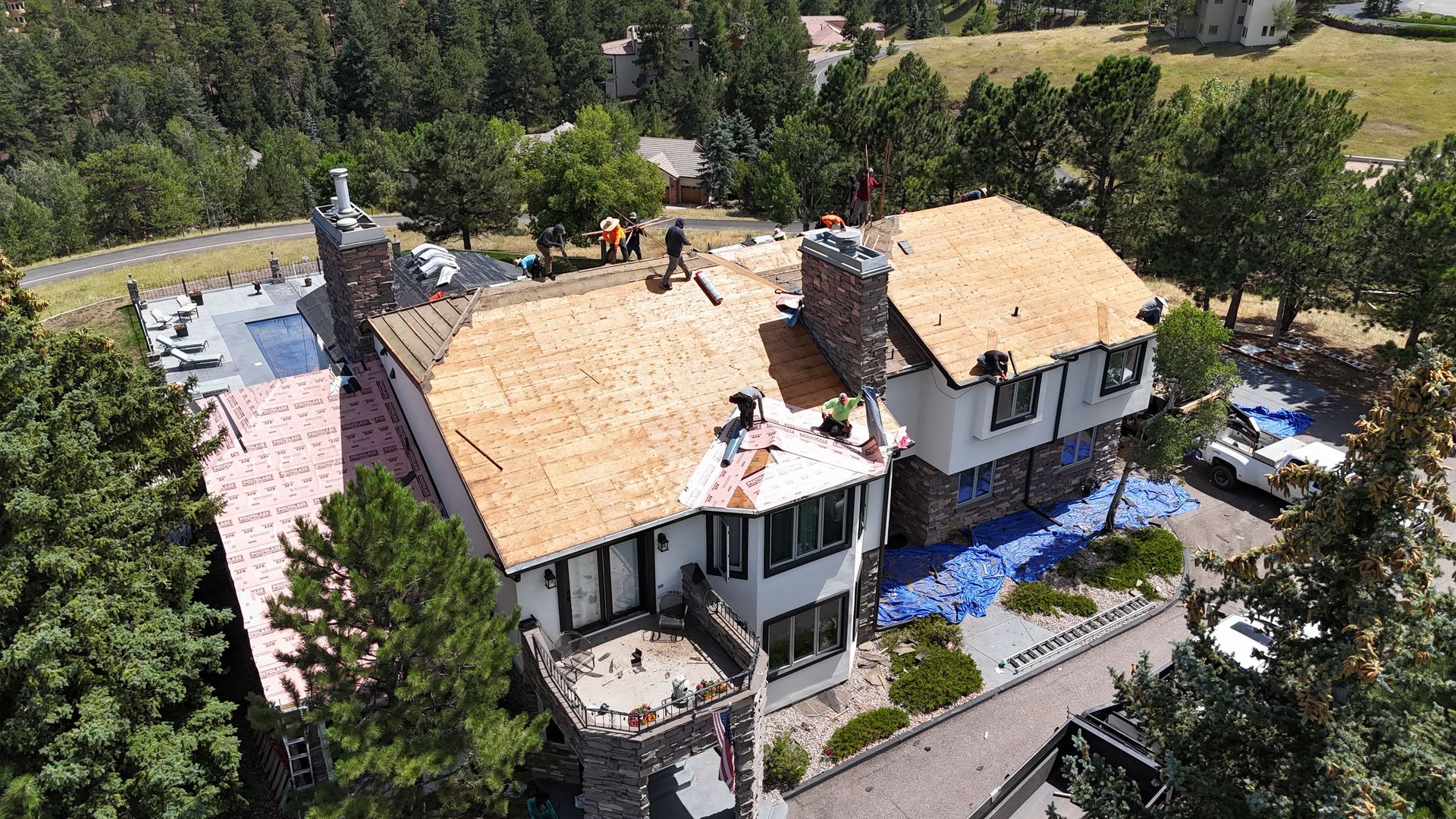 Roofers working on a two-story house with a pool, blue tarps, and surrounding trees.