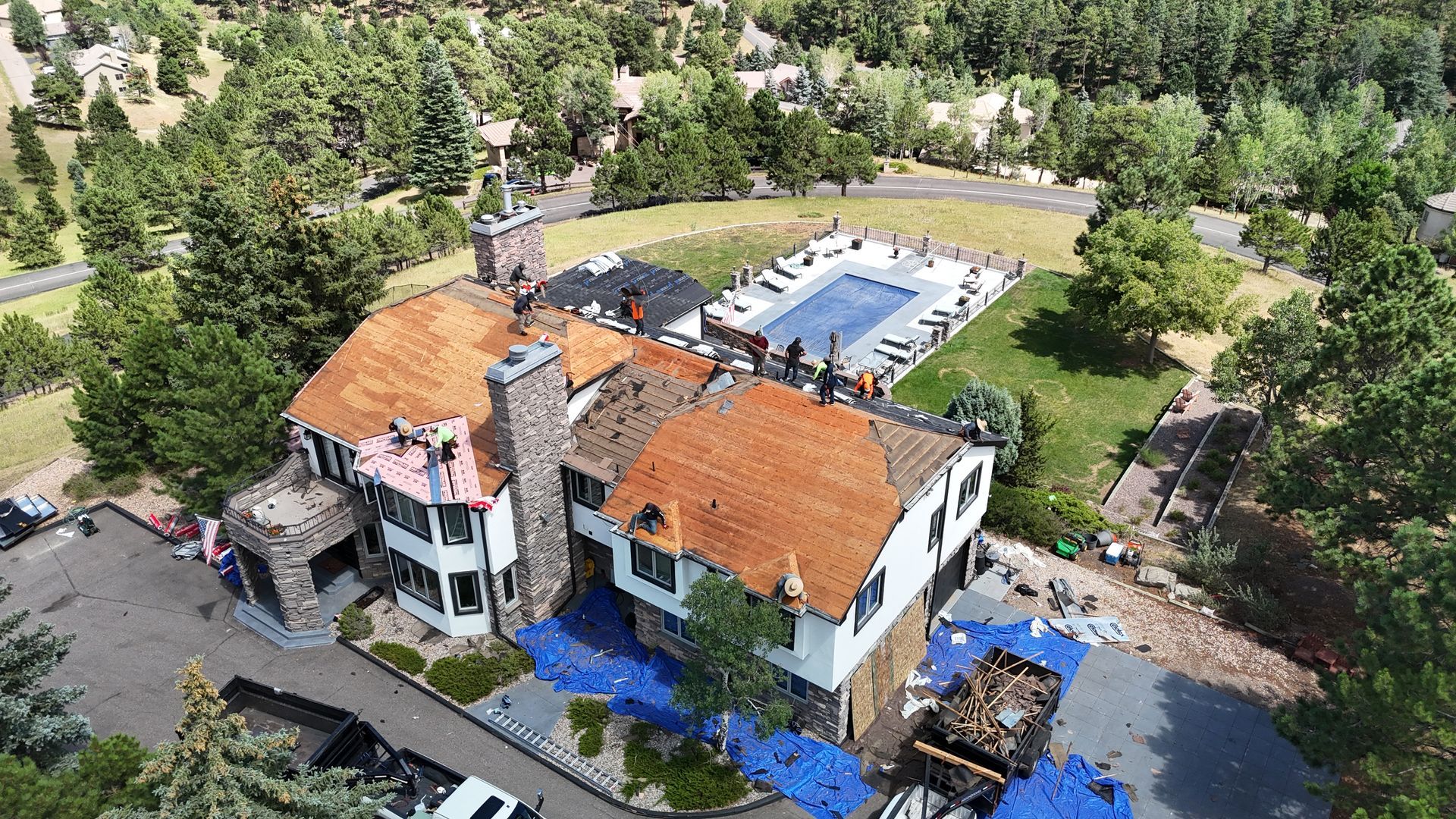 Aerial view of a large house with a roof partially under construction, trees, a pool, and a road in the background.