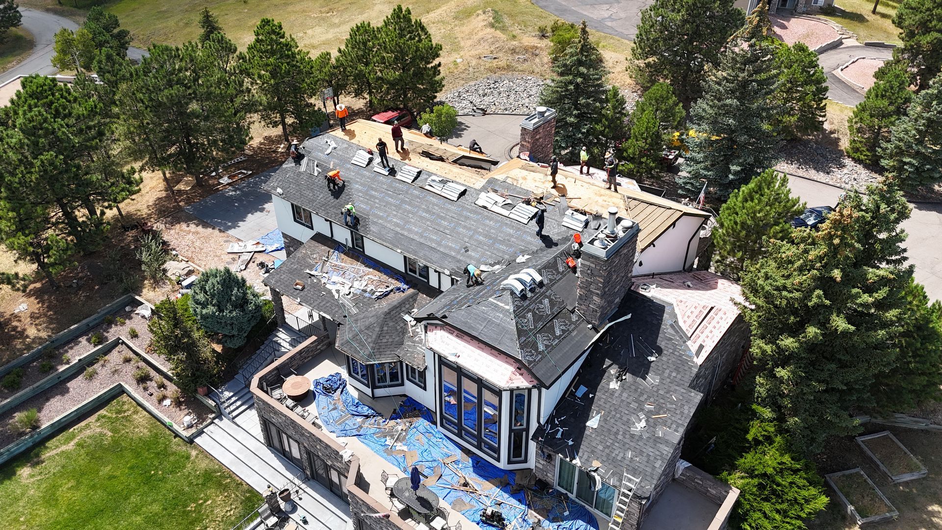 Aerial view: Workers replace a roof on a large house surrounded by trees; blue tarp covers part of the house.