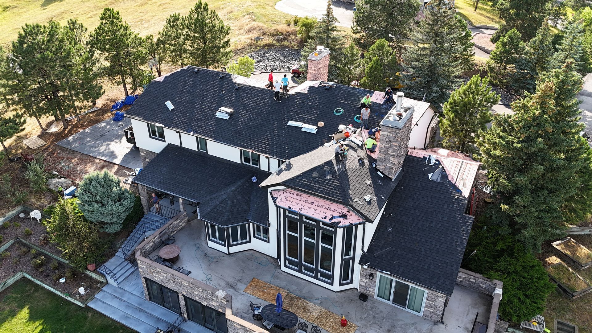 Aerial view of a large house with dark roof, some construction visible. Green trees surround.