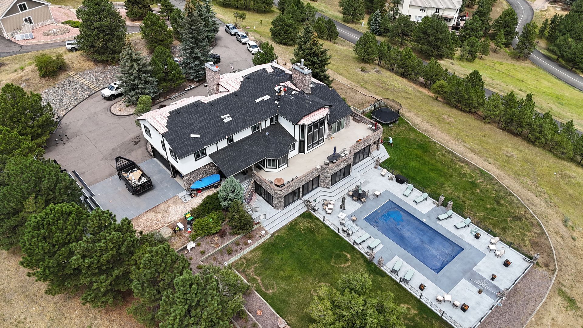 Aerial view of a large house with a pool. The roof is dark, and the surrounding land is green and brown.