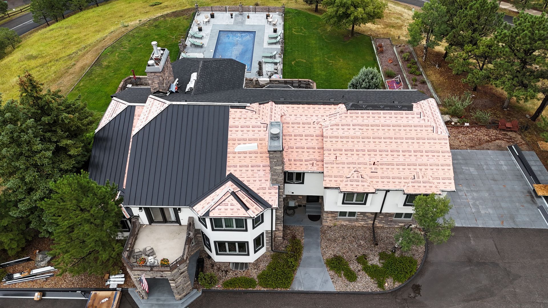 Aerial view of a large mansion with a pool, featuring partially complete roof with contrasting colors and a paved driveway.