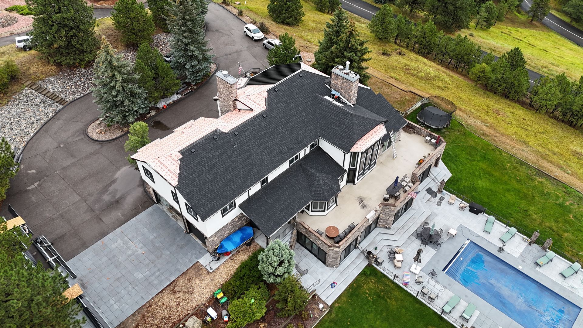 Aerial view of a large house with a pool, driveway, and surrounding trees and grass. The house has a black roof.