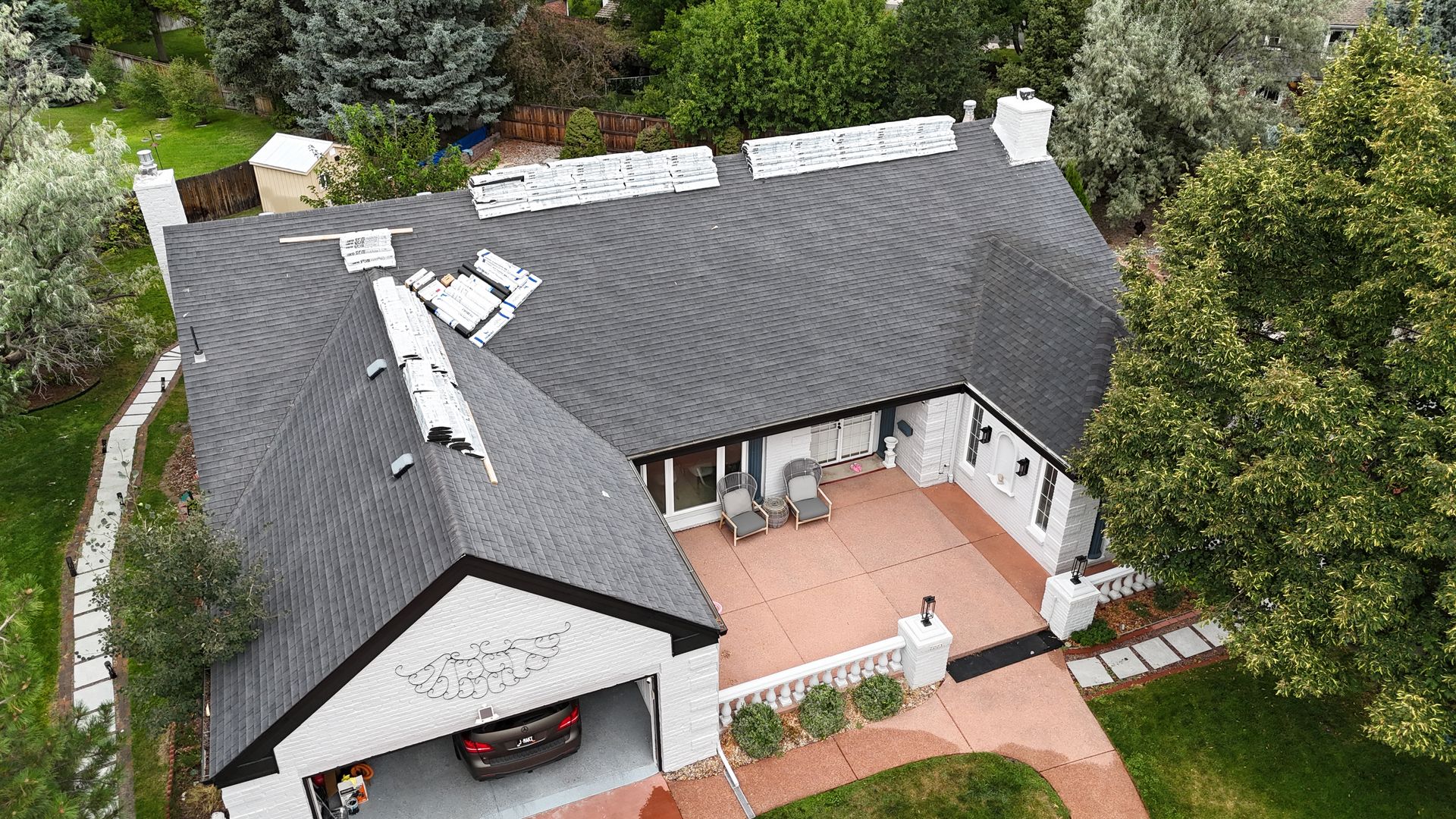Aerial view of a house with a black roof, driveway, and garage. The house is surrounded by green trees.