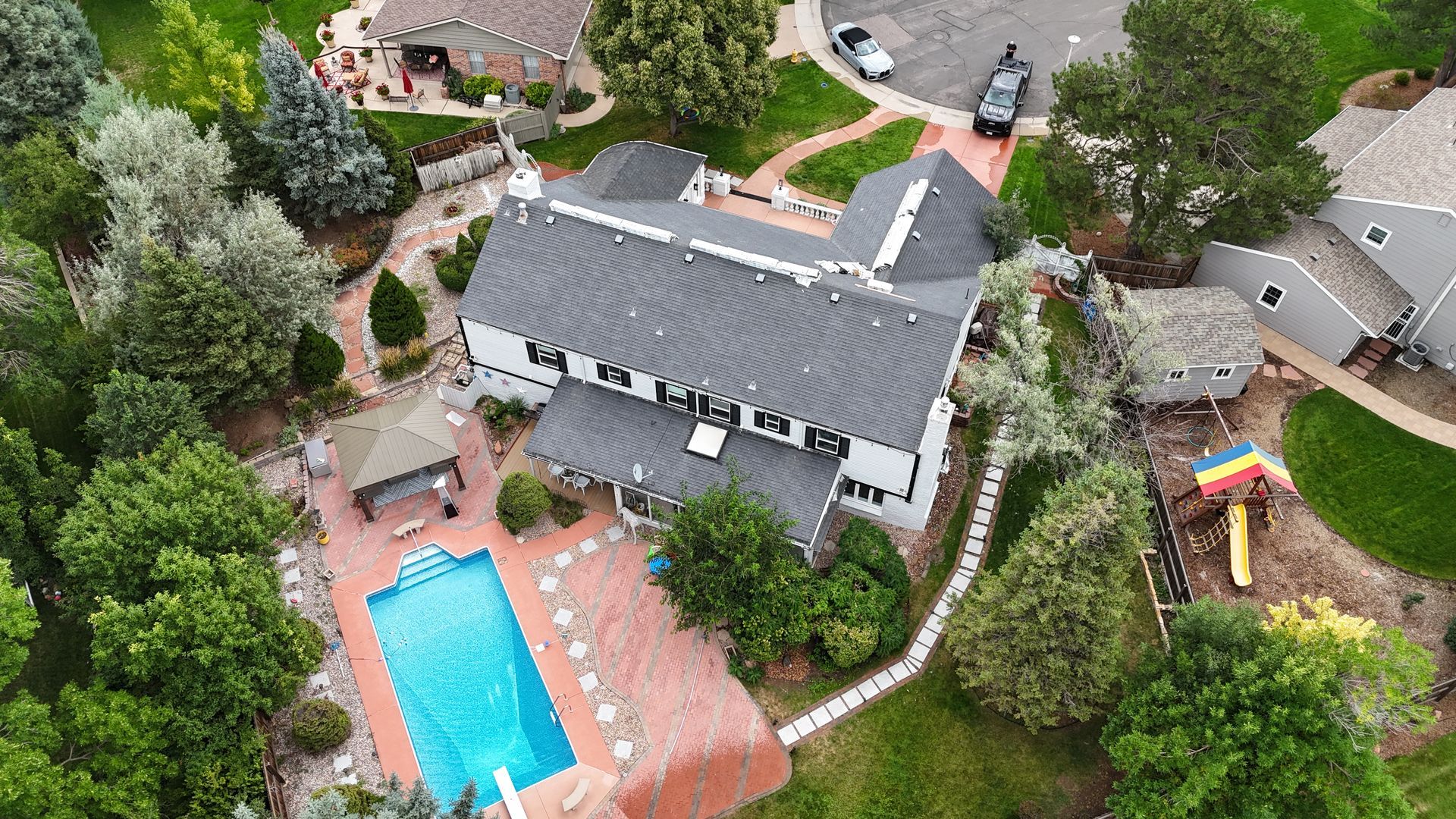 Aerial view of a white house with a pool, surrounded by trees, brick patio, and a small playground.