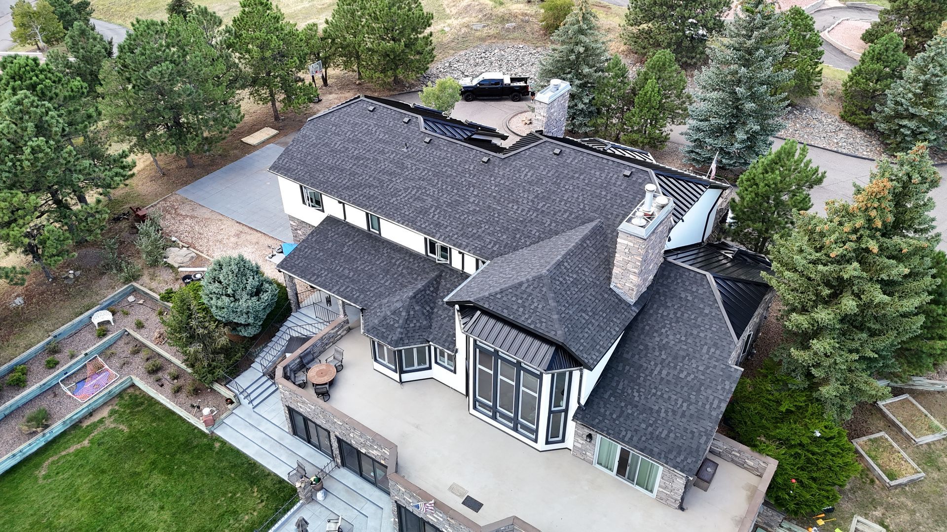 Aerial view of a white and black house with a stone patio, green lawn, and surrounding trees.