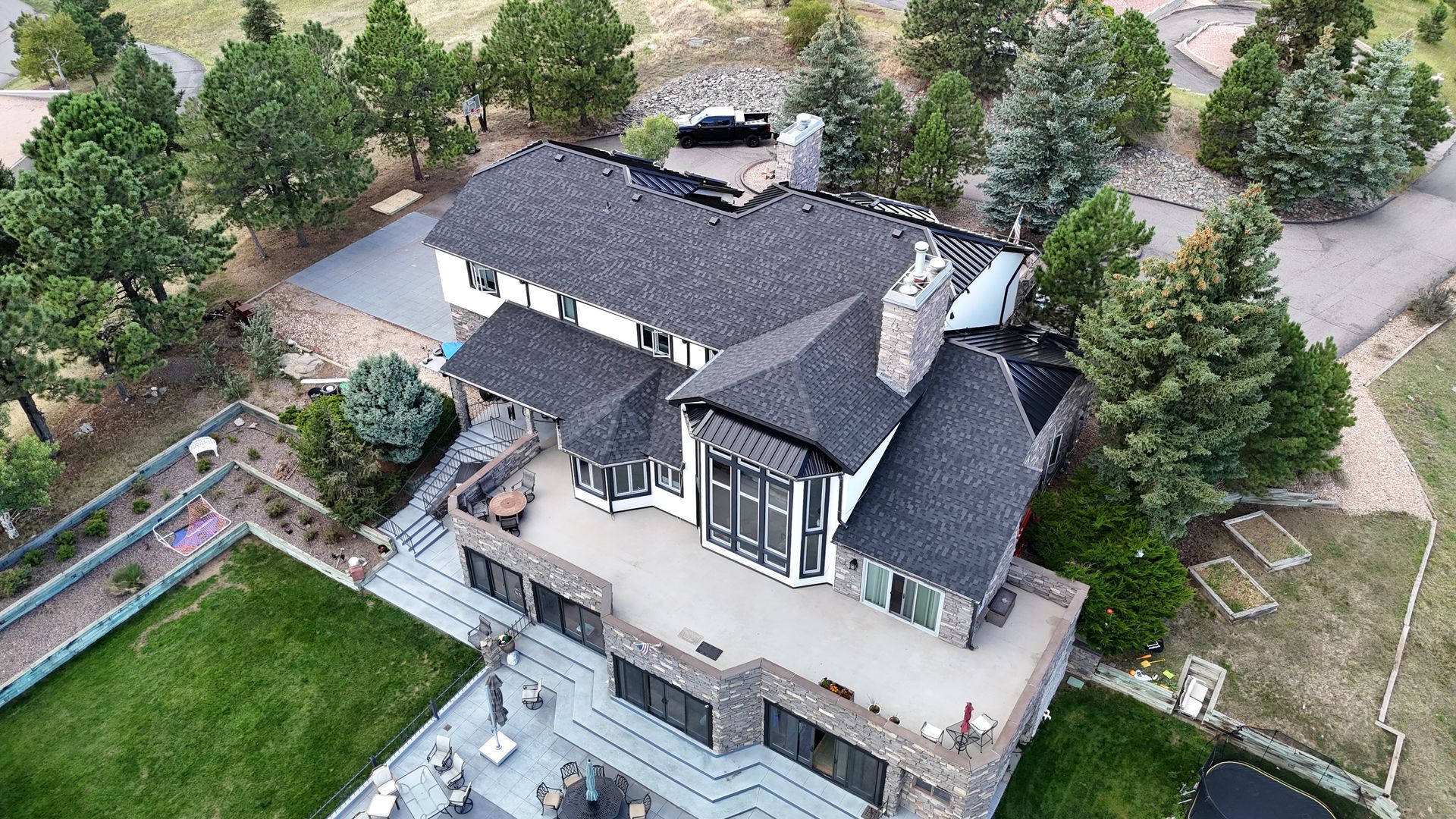 Aerial view of a large, modern house with a gray roof and stone accents surrounded by trees and green lawn.