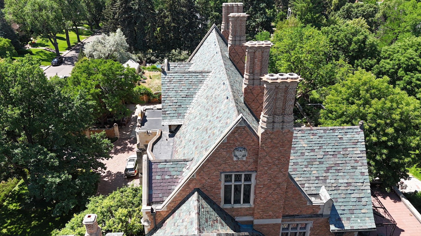 An aerial view of a historic brick mansion with steep slate roofs, multiple chimneys, and surrounding lush green trees.