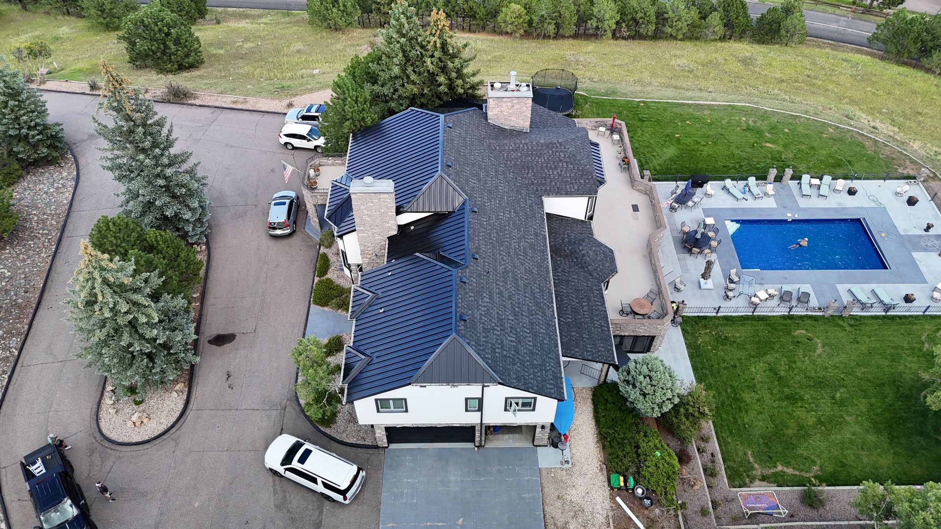Aerial view of a house with a pool, vehicles in the driveway, and surrounding greenery.