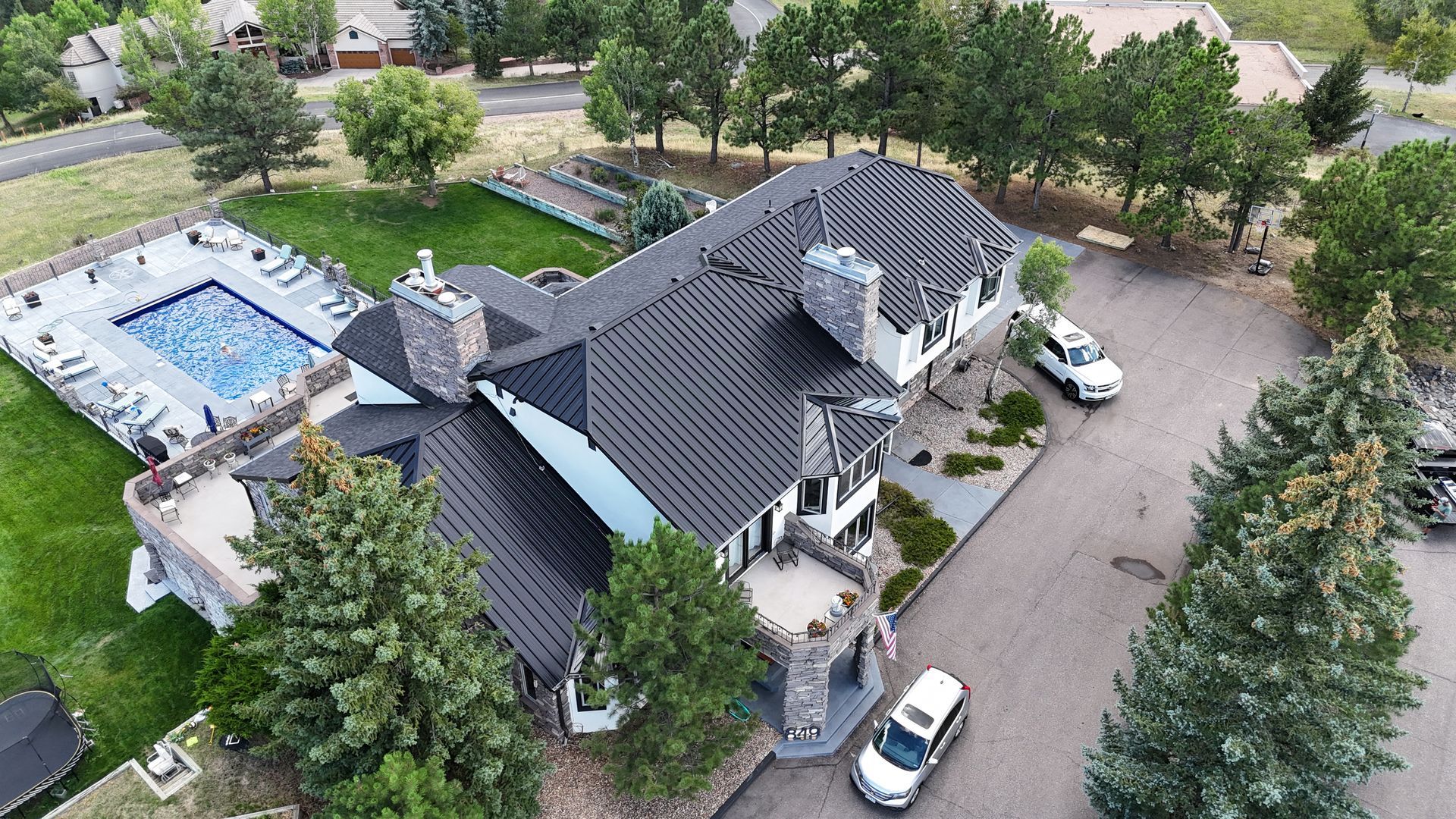 Aerial view of a luxury home with a pool, surrounded by trees, and vehicles parked in the driveway.