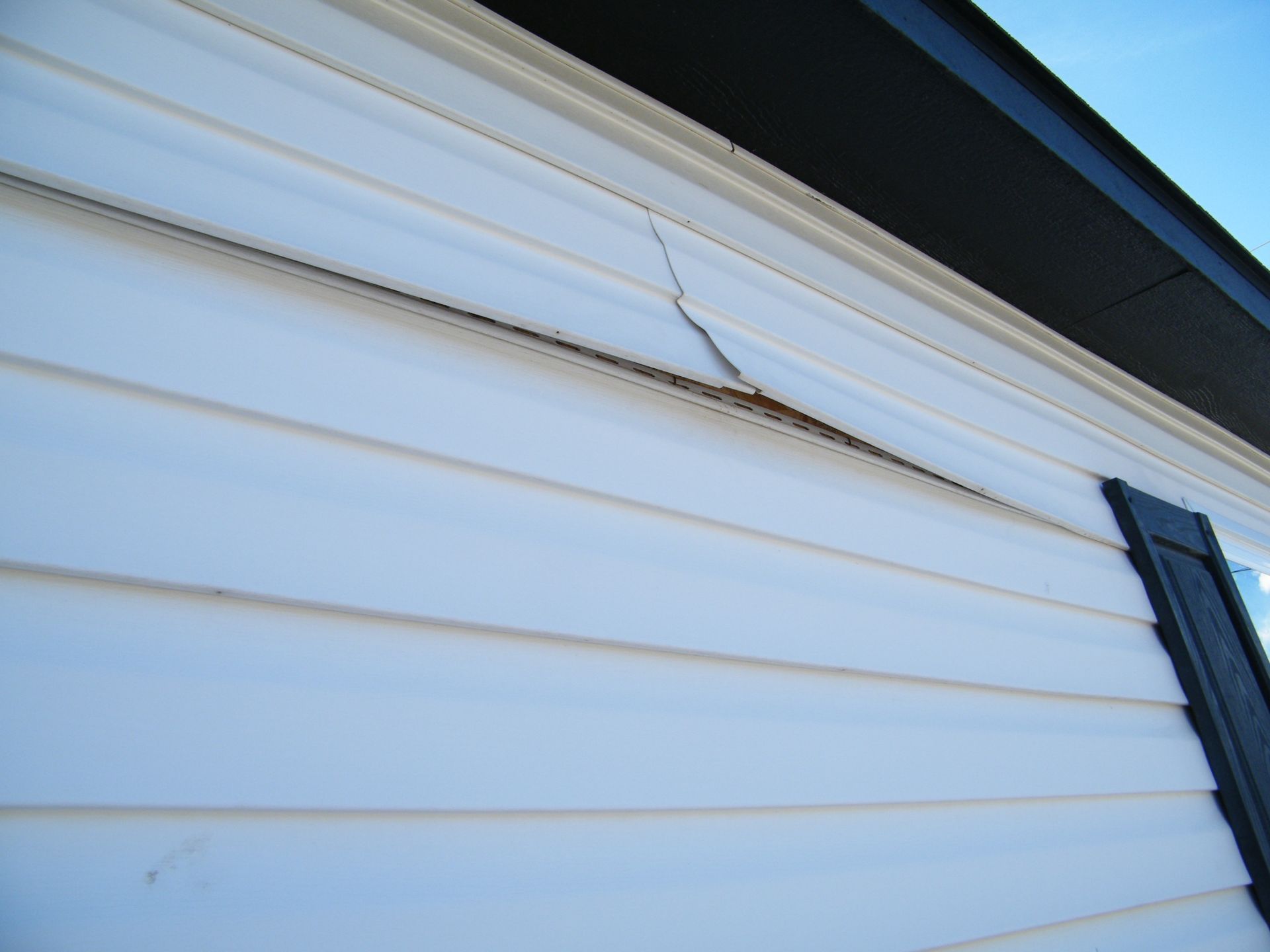 A close-up of white vinyl siding on a house exterior featuring a visible crack and separation near the roofline.