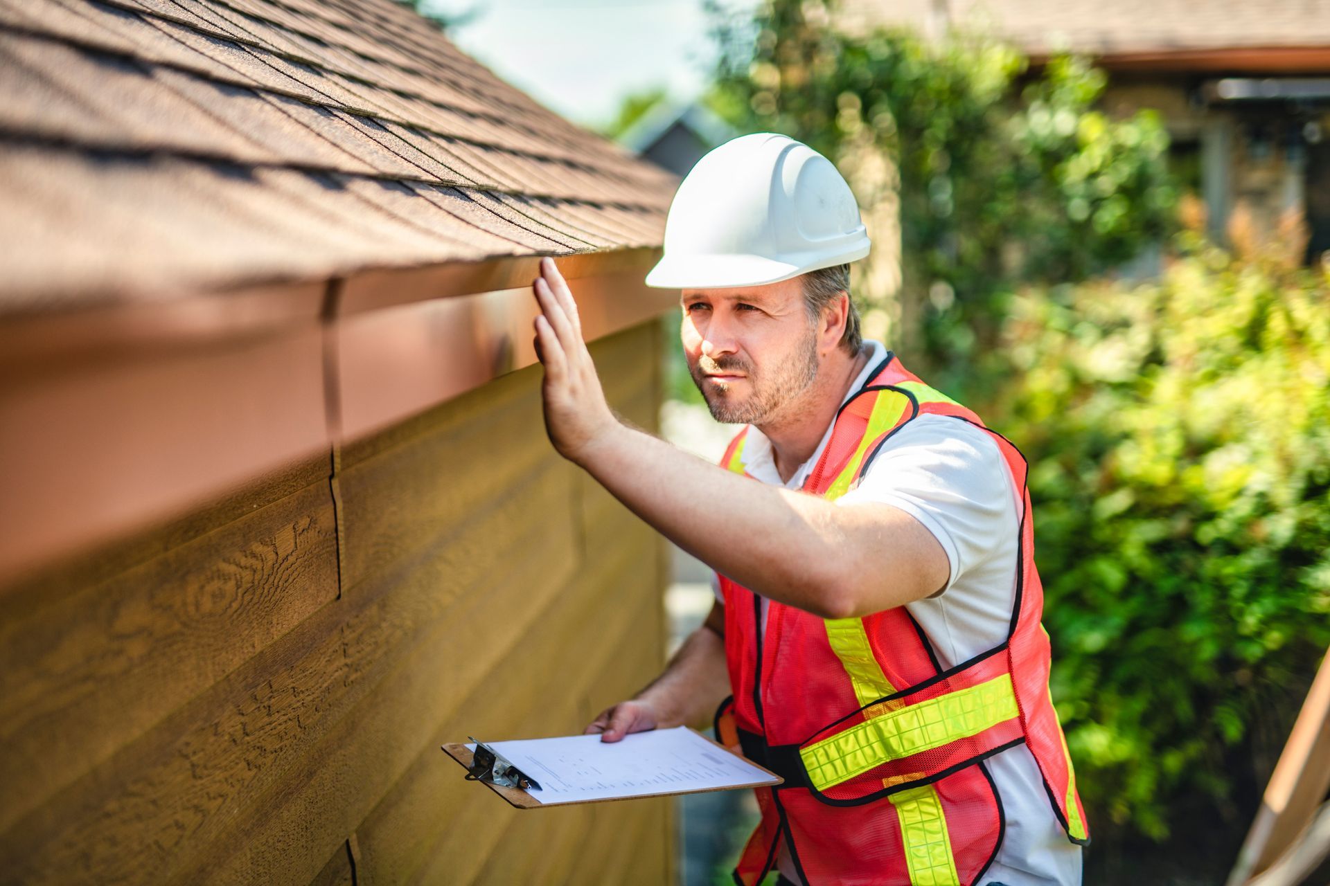 Worker in white hard hat and safety vest inspecting a roof edge with clipboard