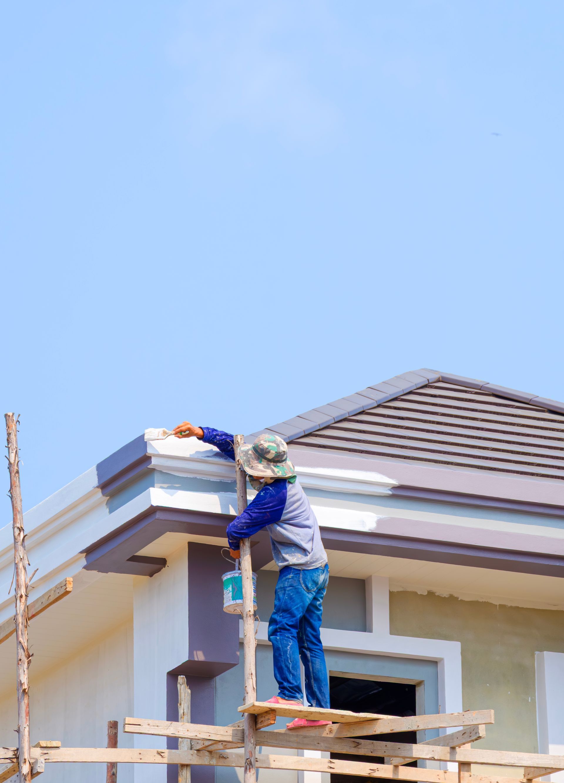 A construction worker on a wooden scaffold painting the exterior trim of a house roof against a clear blue sky.