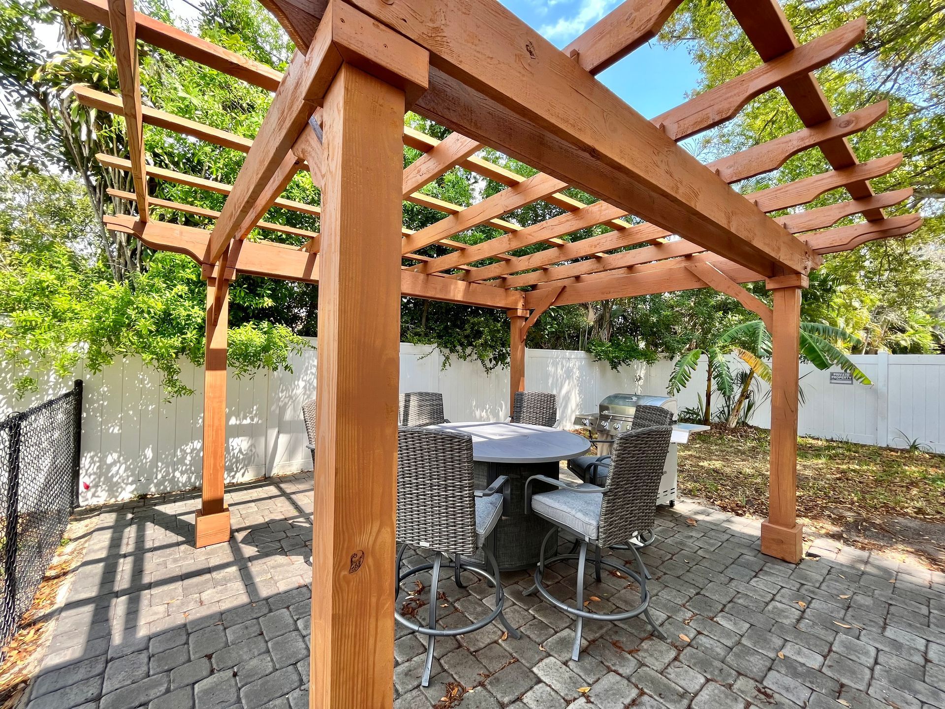 Pergola over patio with table and chairs. Sunny outdoor setting with green foliage and a white fence.