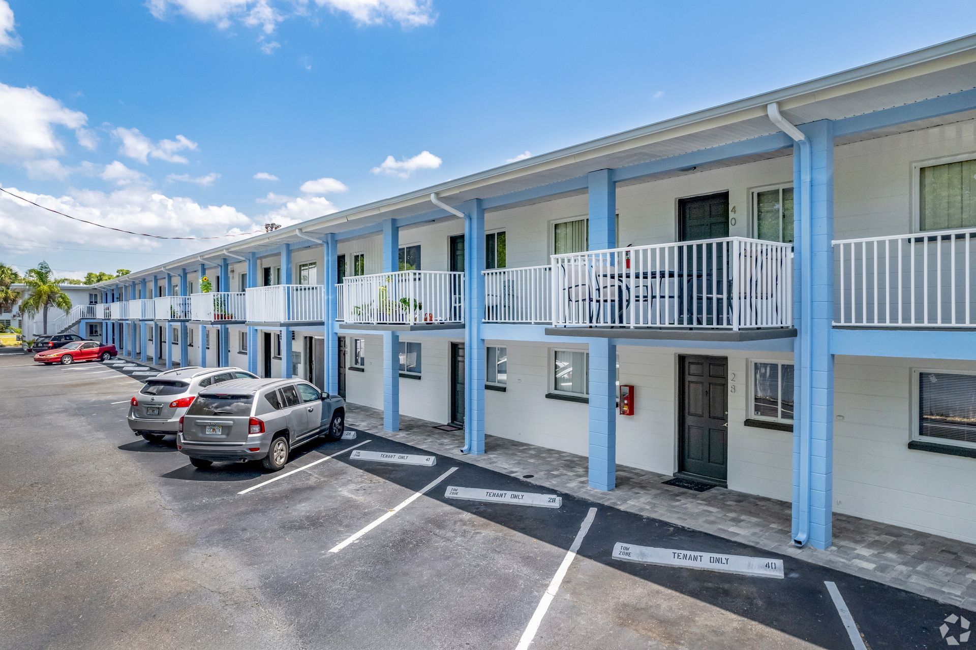 Two-story apartment building with balconies and blue accents; cars parked in front.