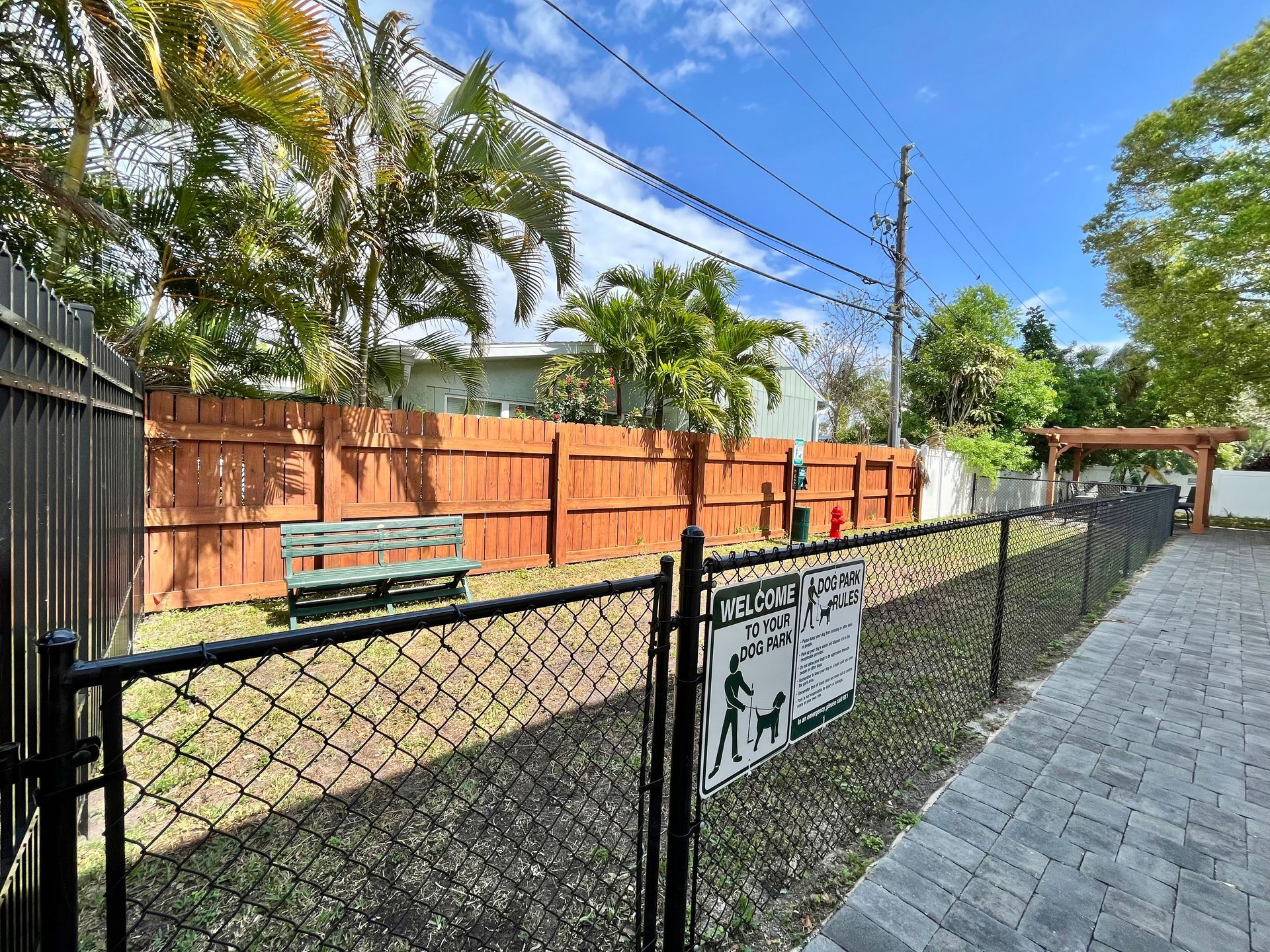 Dog park with chain link fence, wooden bench, and sign. Green grass, brown and green trees, and blue sky.