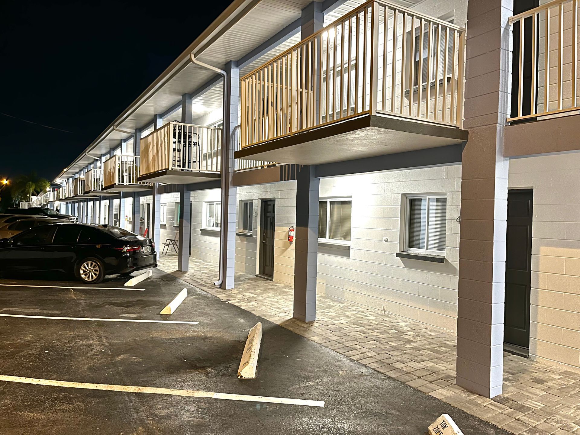 Motel at night. Exterior view shows a row of rooms with balconies, a parking lot, and a car.