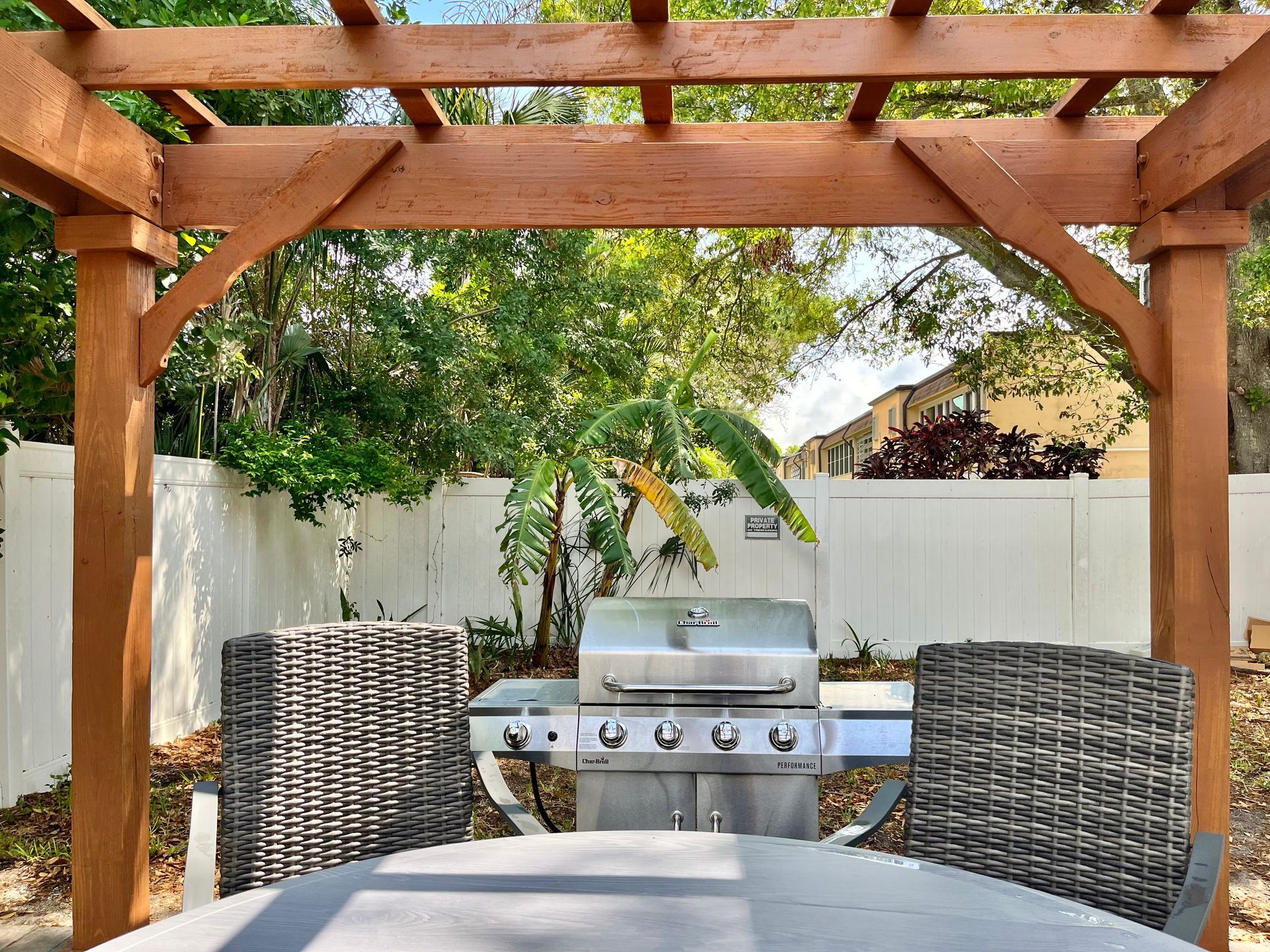 Wooden pergola framing a backyard with a grill between two chairs, and a white fence in the background.