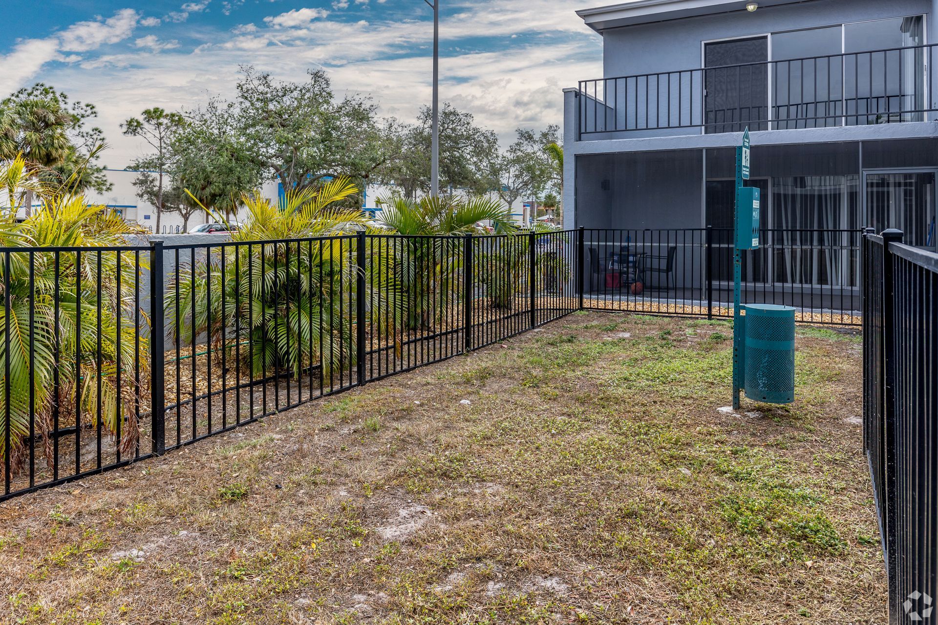 A fenced-in dog park with dry grass, a waste bag dispenser, and a two-story building in the background.