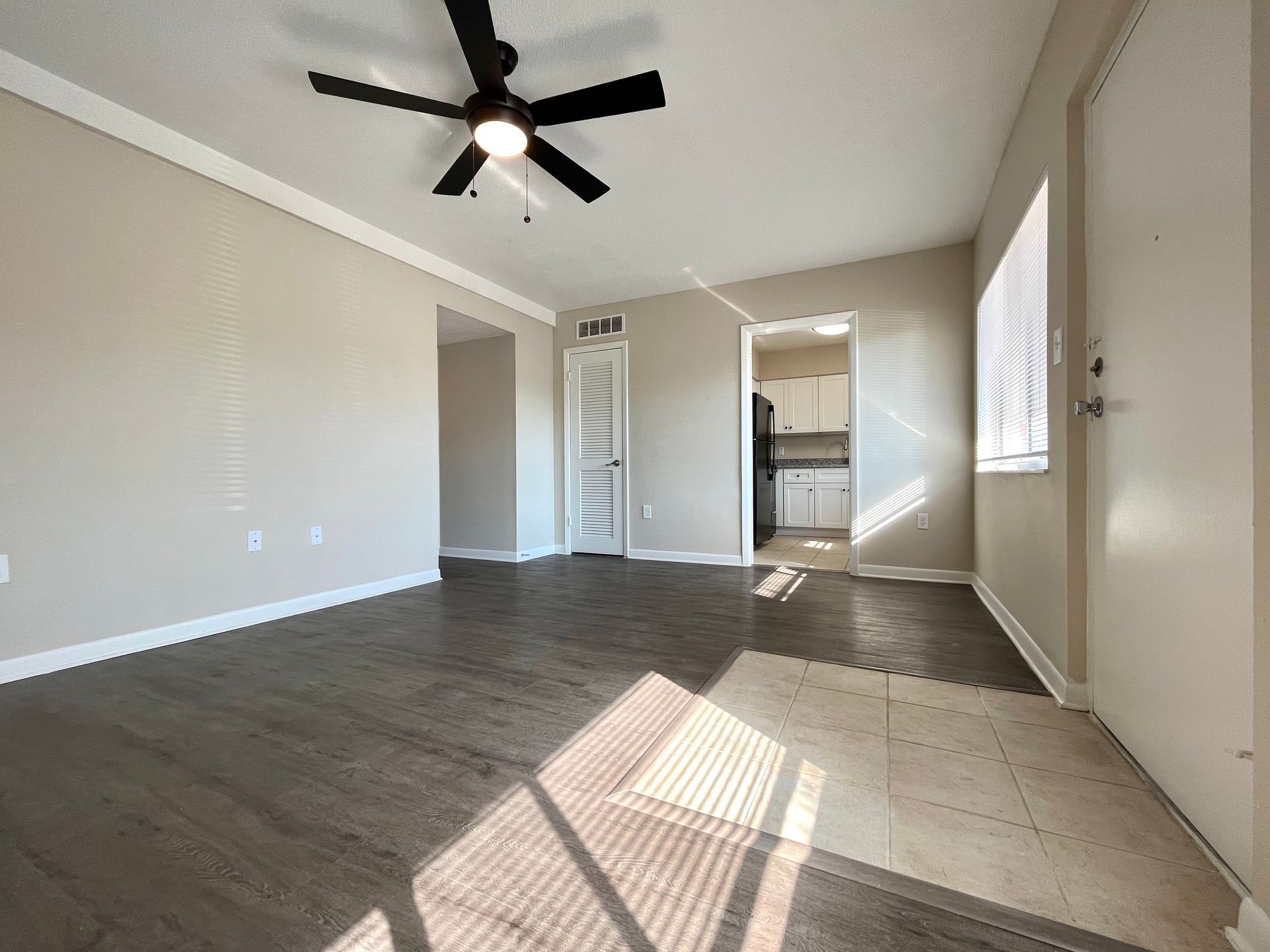 Empty living room with wood-look flooring, beige walls, and a ceiling fan; sunlight streams in from a window.