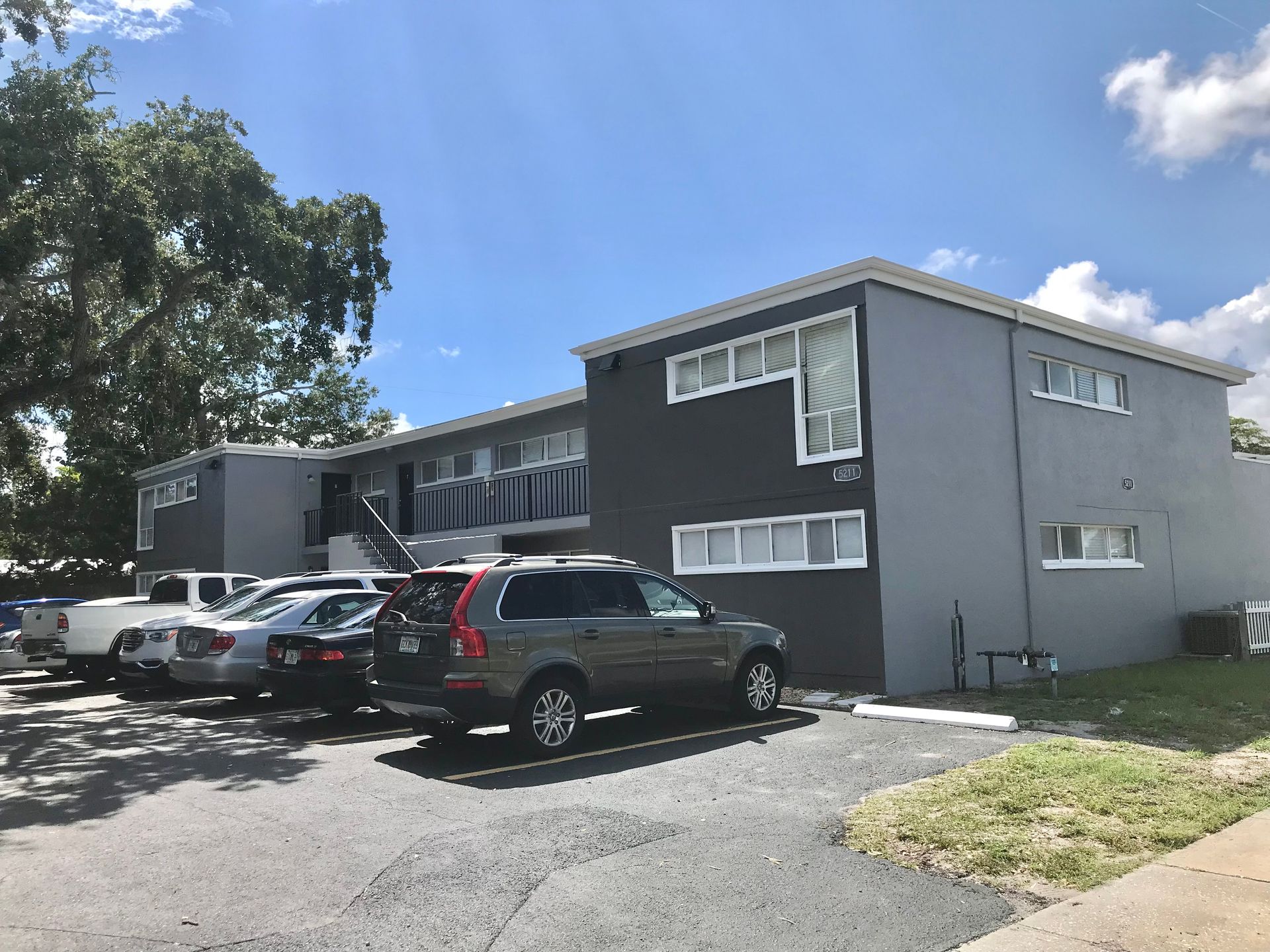 Two-story gray apartment building with cars parked out front on a sunny day.