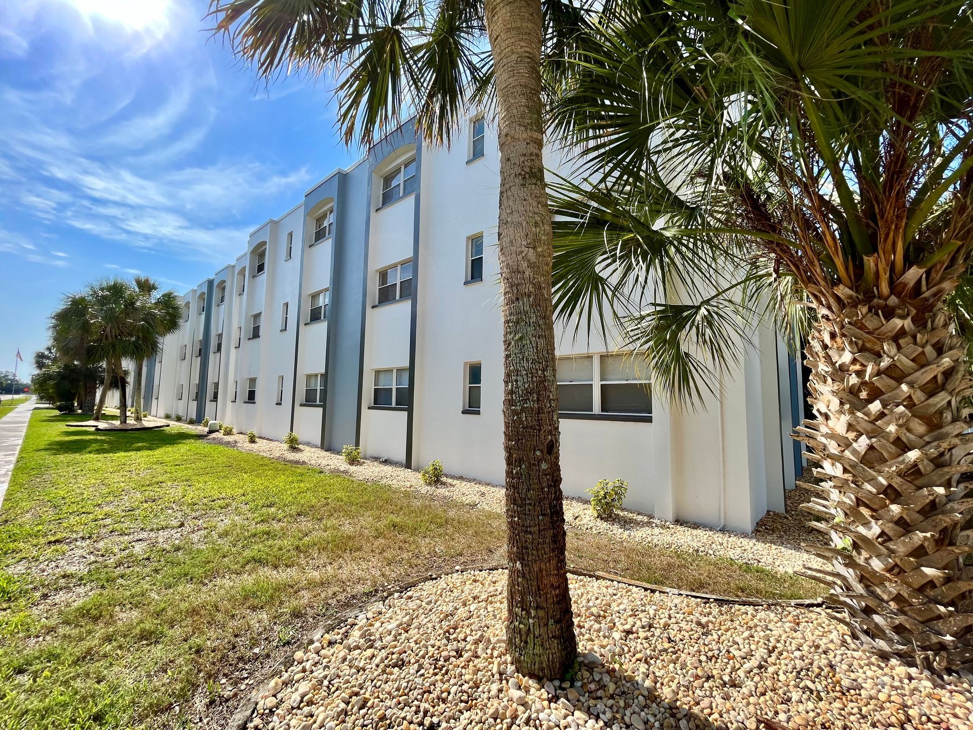 A three-story white building with blue trim sits by a grassy lawn and palm trees on a sunny day.
