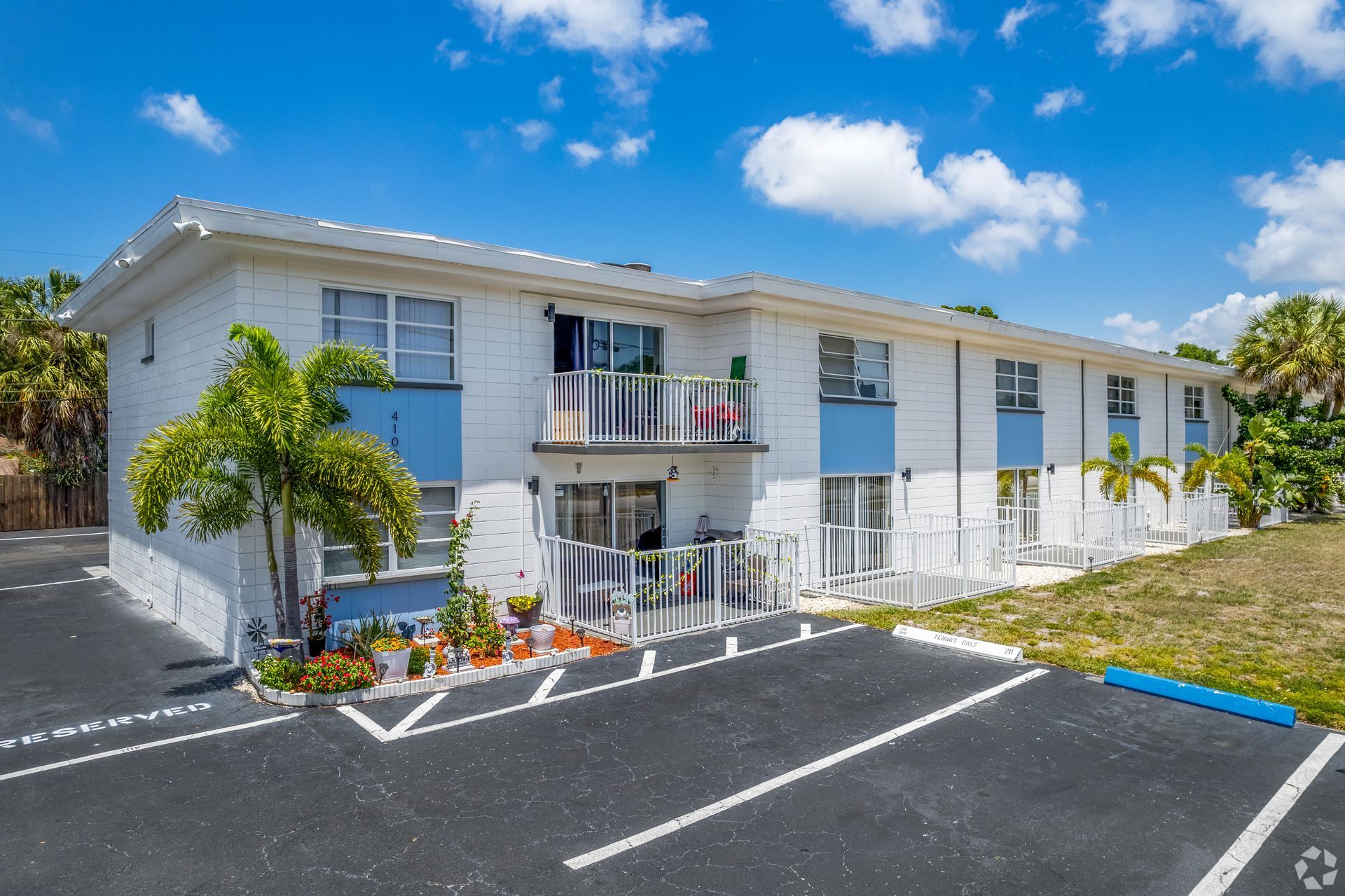 Two-story white and blue apartment building with palm trees and parking spaces on a sunny day.