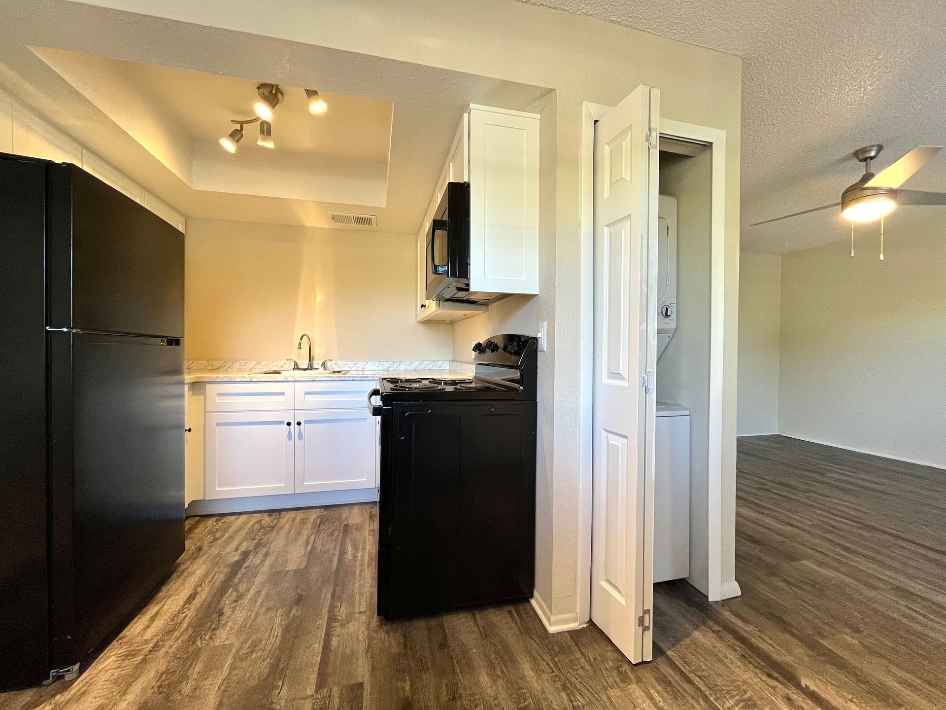 Kitchen with black appliances, white cabinets, and a stacked washer/dryer, near a living area with a ceiling fan.