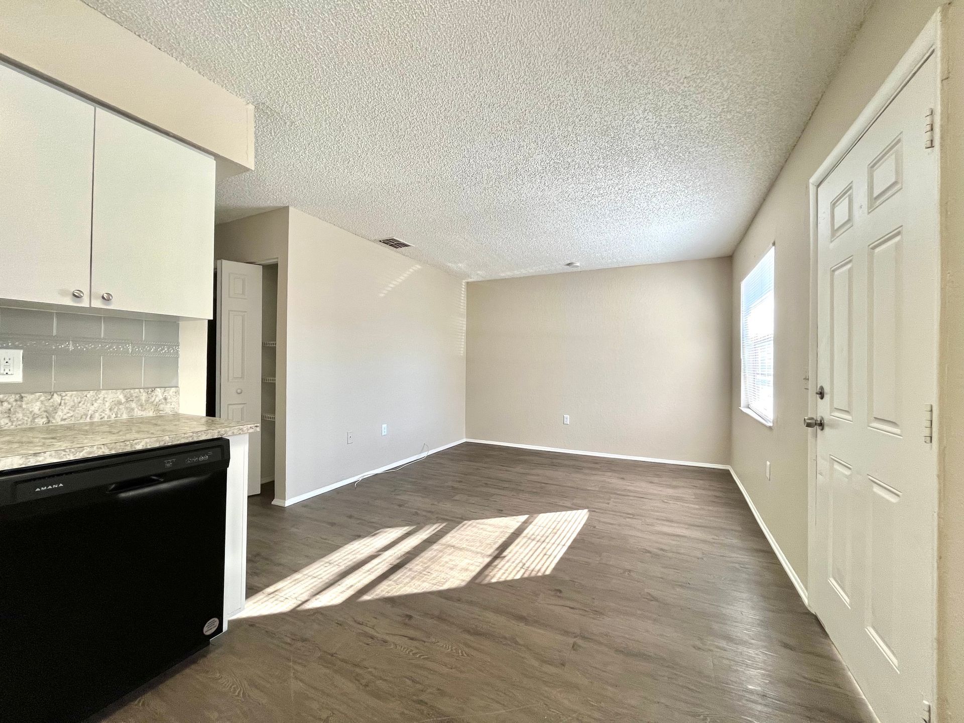 Empty living room with kitchen access, tan walls, wood-look floor, white door, and black appliances.