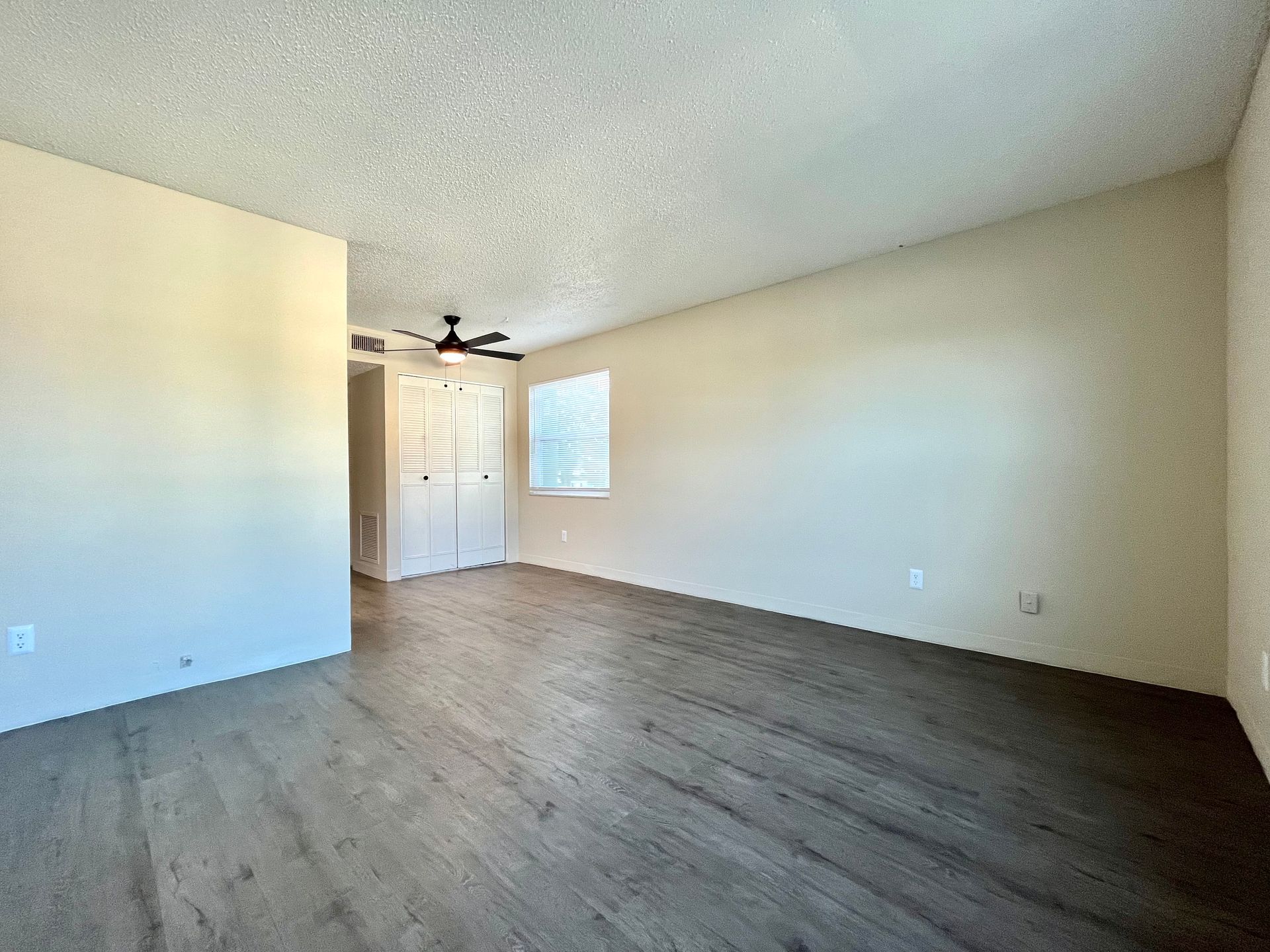 Empty room with light wood-look flooring, off-white walls and popcorn ceiling. Light comes from a window.
