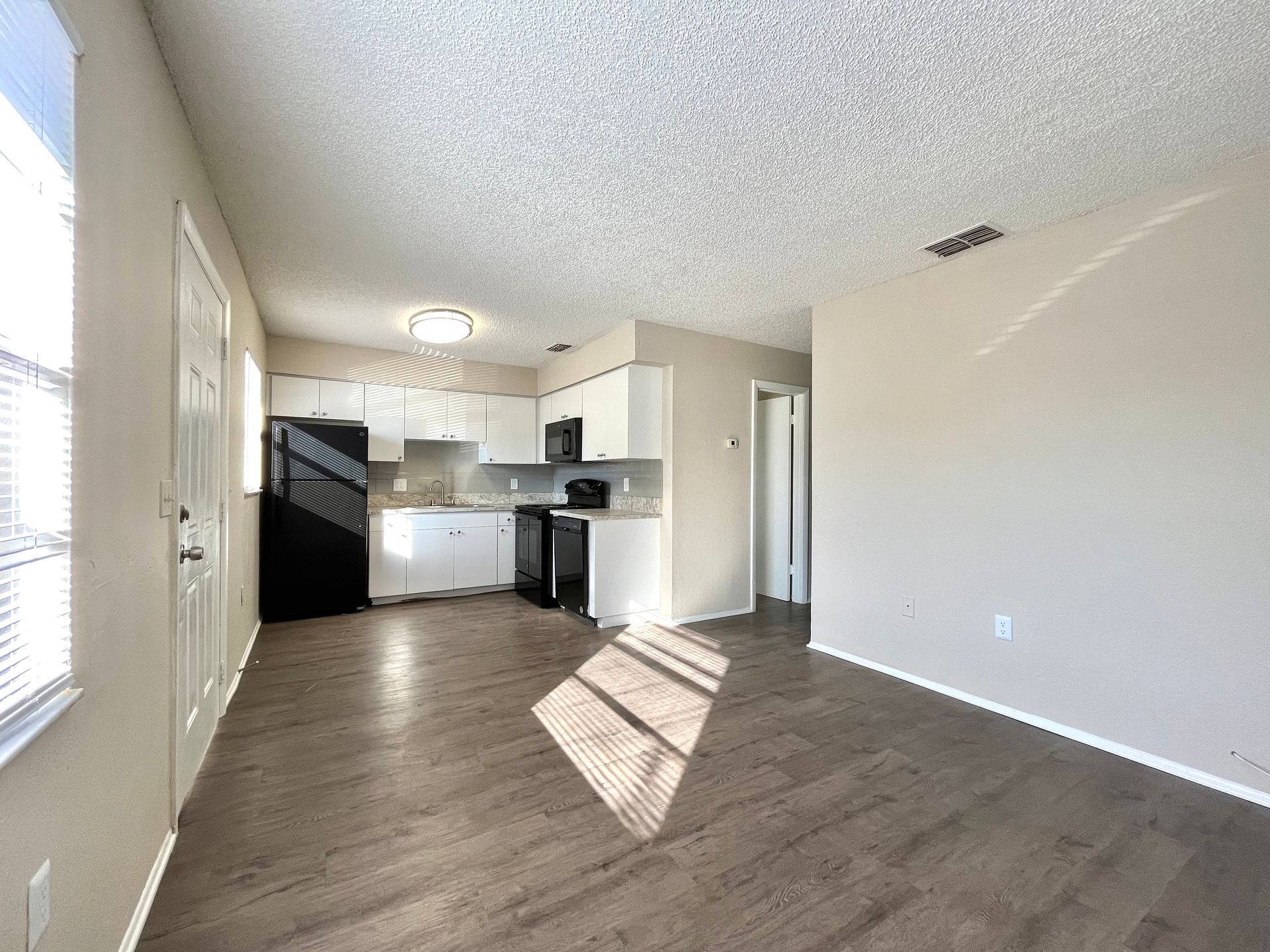 Interior view of a kitchen and living area with white cabinets, dark appliances, and wood-look flooring.
