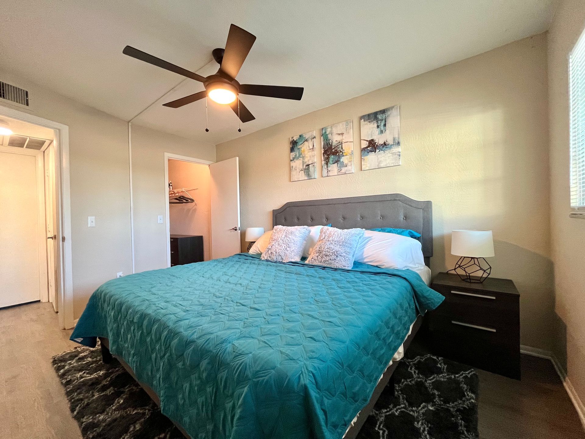 Bedroom with teal bedspread, gray headboard, and artwork above the bed. A ceiling fan hangs overhead.
